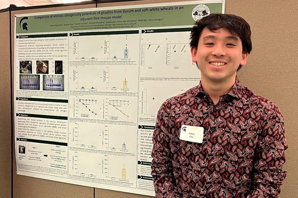 A man with short black hair who is smiling and is wearing a nametag that says "Jaden Loy.' He is standing next to a research poster.