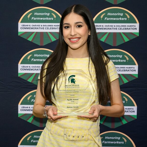 A young woman in a yellow dress smiles and holds a clear glass award with the Michigan State University logo in front of a backdrop that reads “Honoring Farmworkers: César E. Chávez & Dolores Huerta Commemorative Celebration.”
