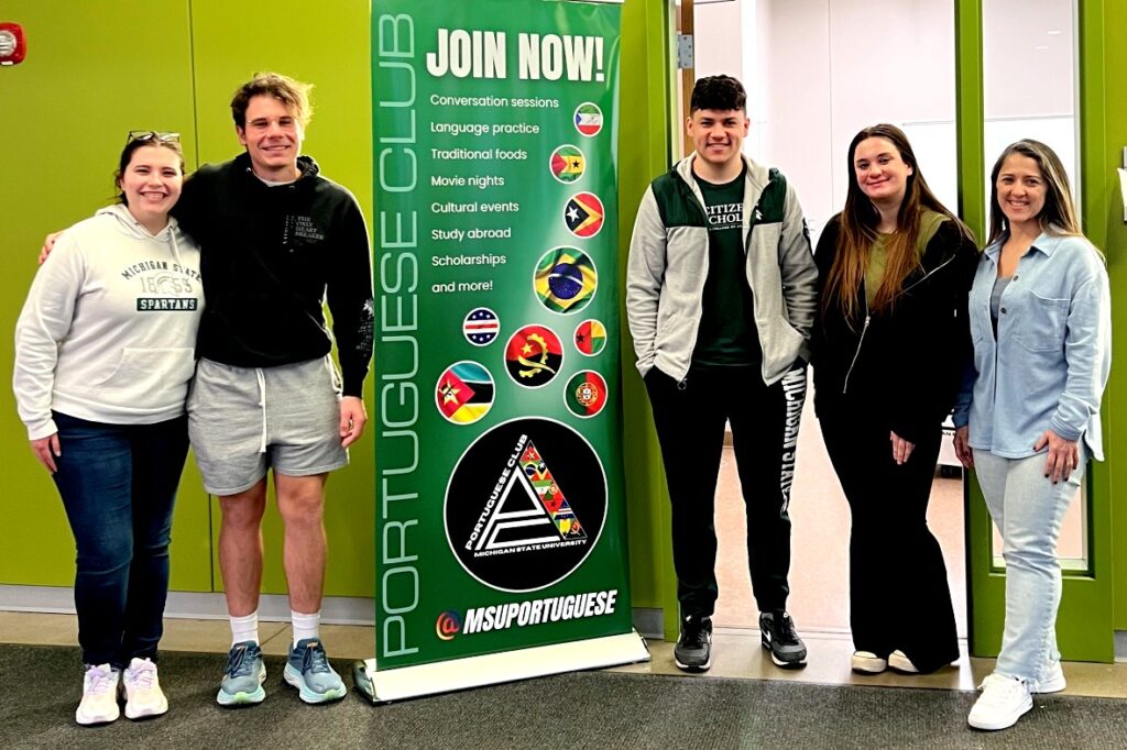 Five people stand in front of a green banner promoting the MSU Portuguese Club, smiling; the banner lists activities like conversation sessions, study abroad, and scholarships.