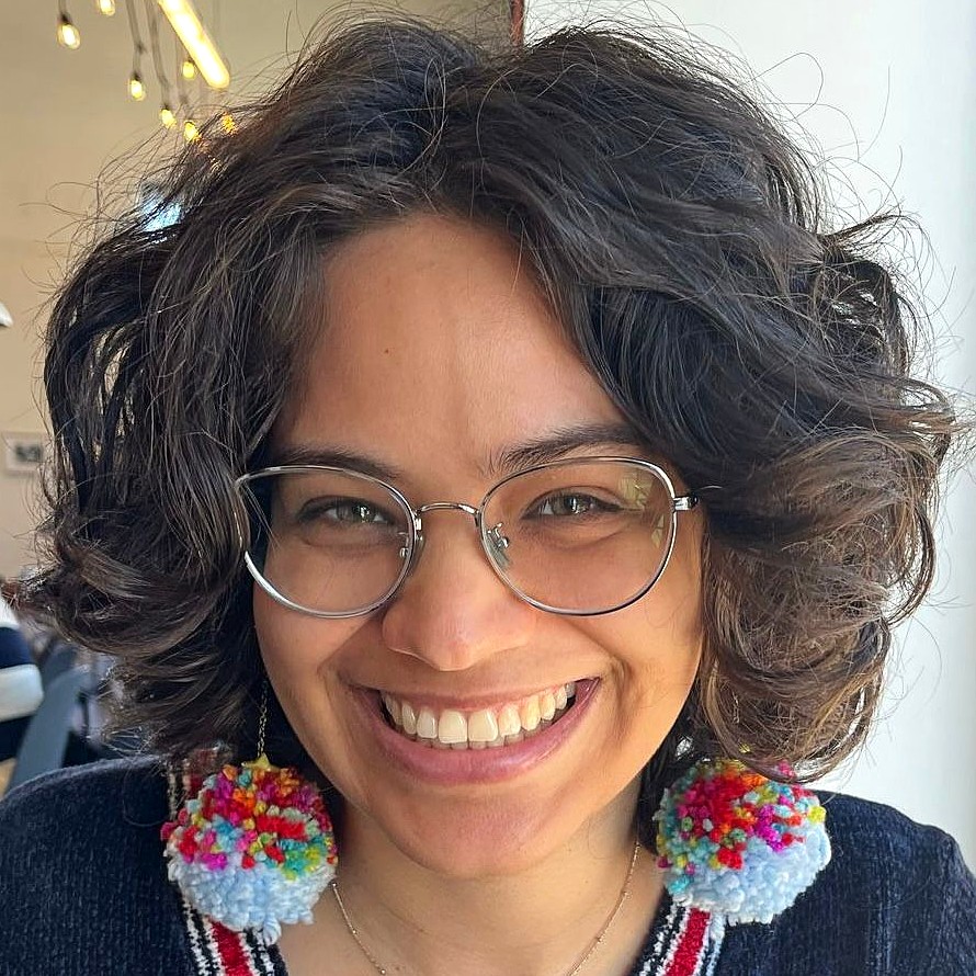 Woman with should-length black hair, smiling, wearing glasses with a silver frame and wearing a multi-colored pom-pom earrings.