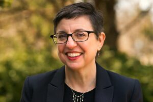 A woman with short dark hair and glasses smiles outdoors. She is wearing a dark blazer, a black top, drop earrings, and a silver necklace. The background is filled with greenery and trees.