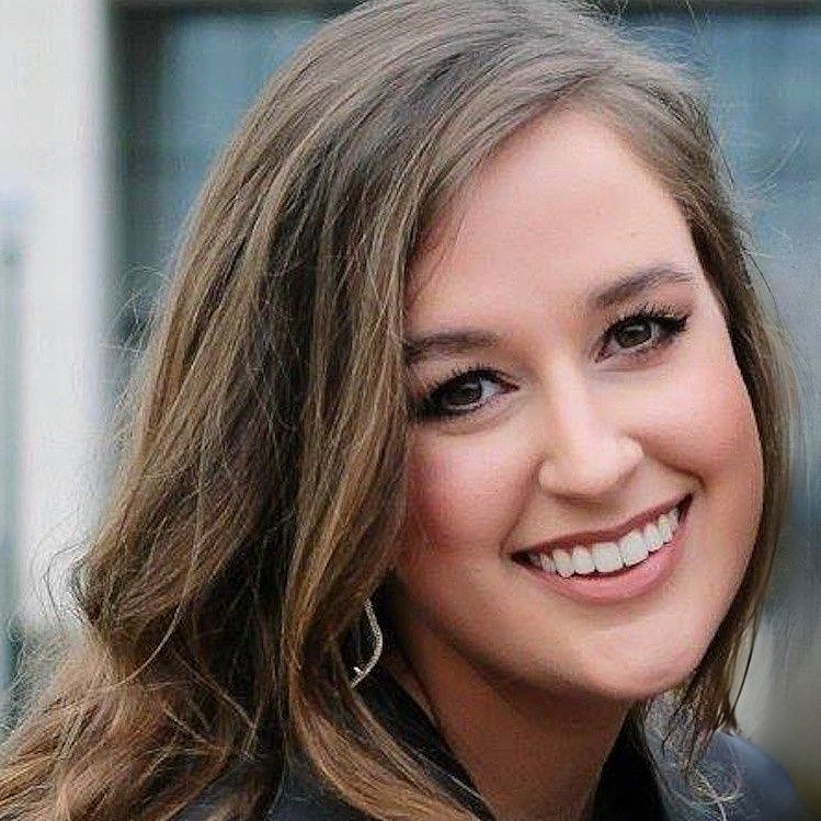 A picture of a woman with long brown hair and brown eyes smiling iwith her head tilted to one side.