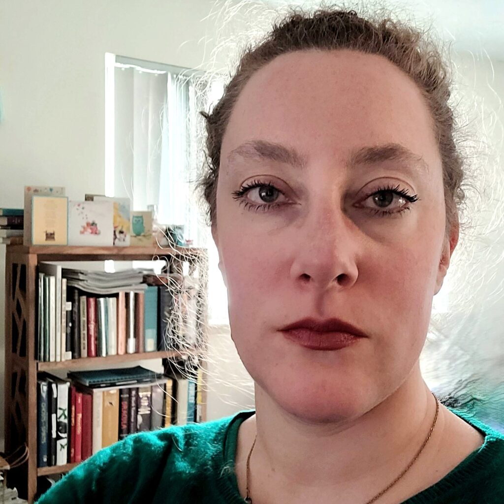 A picture of a woman in a green shirt looking at the camera. Bookshelves are visible behind her.