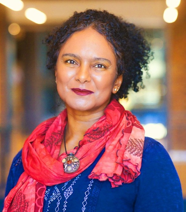 Headshot of a woman with black curly hair pulled up, with dark red lipstick, a red scarf and a blue shirt. 