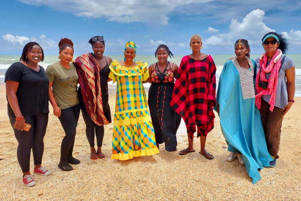 A group of eight Black women in brightly colored clothes standing in a row on a beach. 