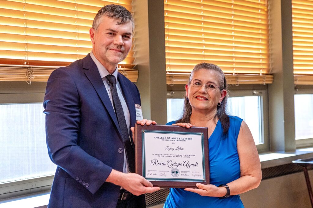 A man in a navy suit and tie stands next to a woman in a sleeveless blue blouse. Both are smiling while holding a framed award together. The plaque reads, “Legacy Lecture – Rocío Quispe-Agnoli, College of Arts & Letters, Michigan State University.”