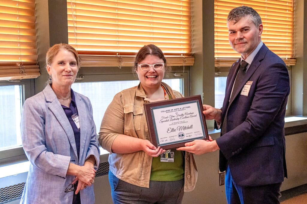 Three people stand together in a room with windows and blinds. The person in the center, wearing glasses and a tan jacket, smiles while holding a framed plaque. On the left stands a woman in a light purple blazer, and on the right a man in a navy suit and tie holds the other side of the plaque. The award reads, “Fixed-Term Faculty/Academic Specialist Leadership Excellence Award – Ellie Mitchell, College of Arts & Letters, Michigan State University.”