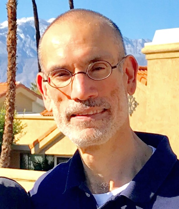 A man with short hair, a graying beard, and round glasses stands outdoors in bright sunlight. Behind him are tan stucco buildings, tall palm trees, and snow-capped mountains.
