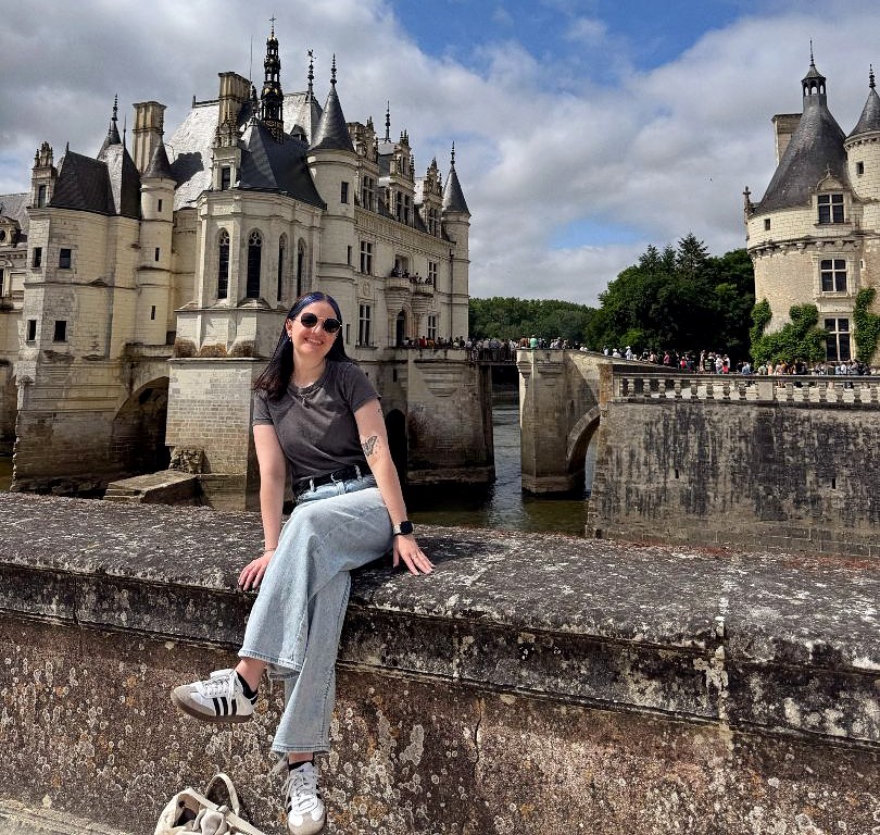 Womn sitting on a cement ledge next to a river with a large castle in the background. 