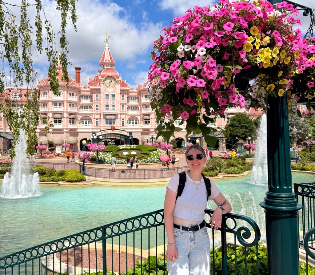 Woman standing next to a pool of water with a fountain. There is a large pink flowering plant above her head and a beautiful pink building in the background.  