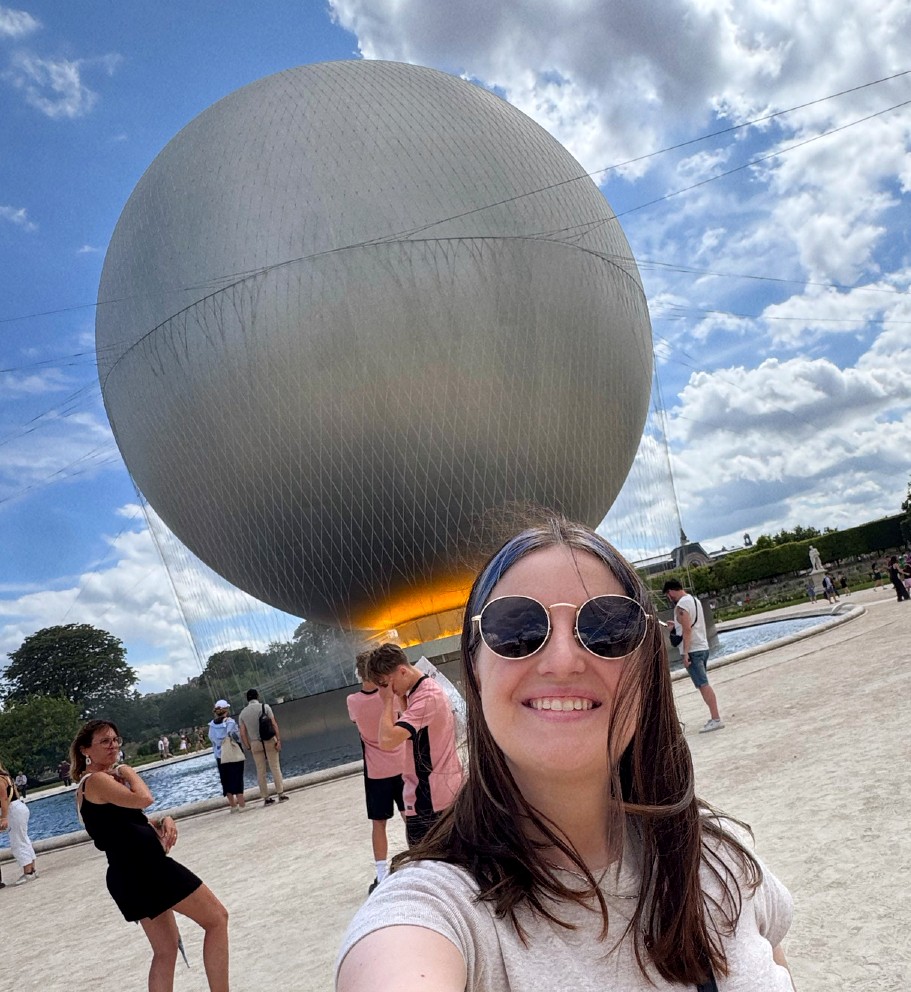 a selfie photo of a woman with dark long hair and dark sunglasses. There is a large sphere behind her that appears to be lit up at the bottom. 