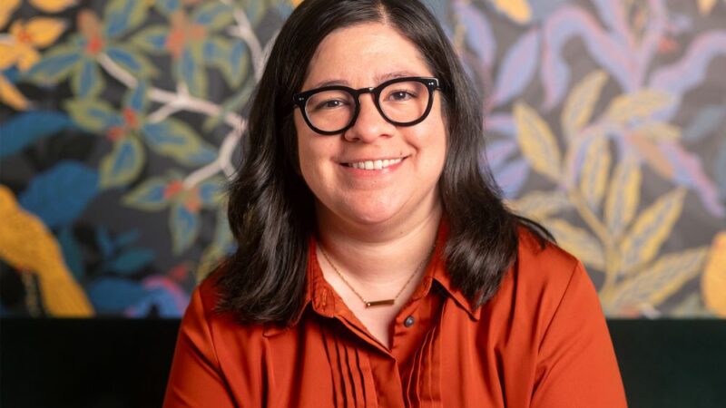 Alejandra Márquez, Assistant Professor of Spanish at Michigan State University, wears an orange shirt and glasses. She sits in from a wall with wallpaper that shows green, blue, and yellows leaves.