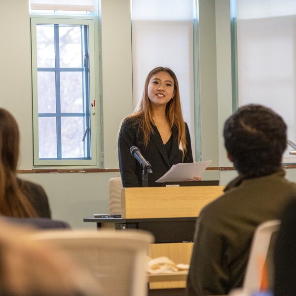 Rachel Zhai stands at a podium holding notes while delivering an oral presentation to an audience seated in a lecture-style room at the Diversity Research Showcase.