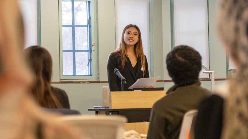 Rachel Zhai stands at a podium holding notes while delivering an oral presentation to an audience seated in a lecture-style room at the Diversity Research Showcase.