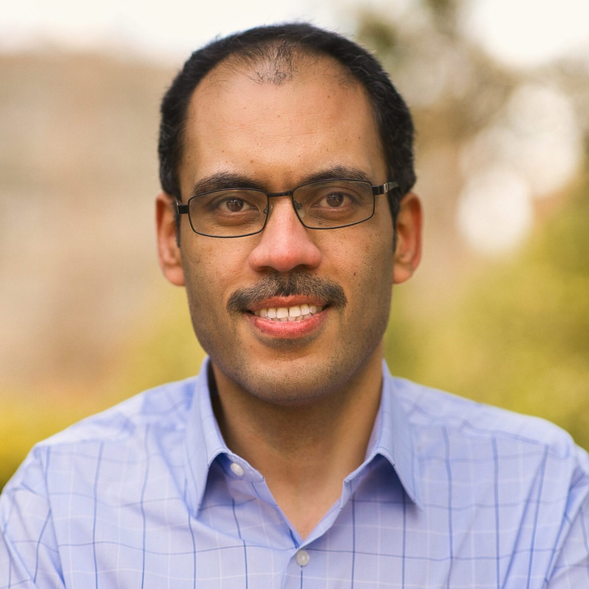 Professional headshot of Ayman Mohamed wearing glasses and a light blue checkered button-down shirt. He is smiling against a soft-focus, golden outdoor background.