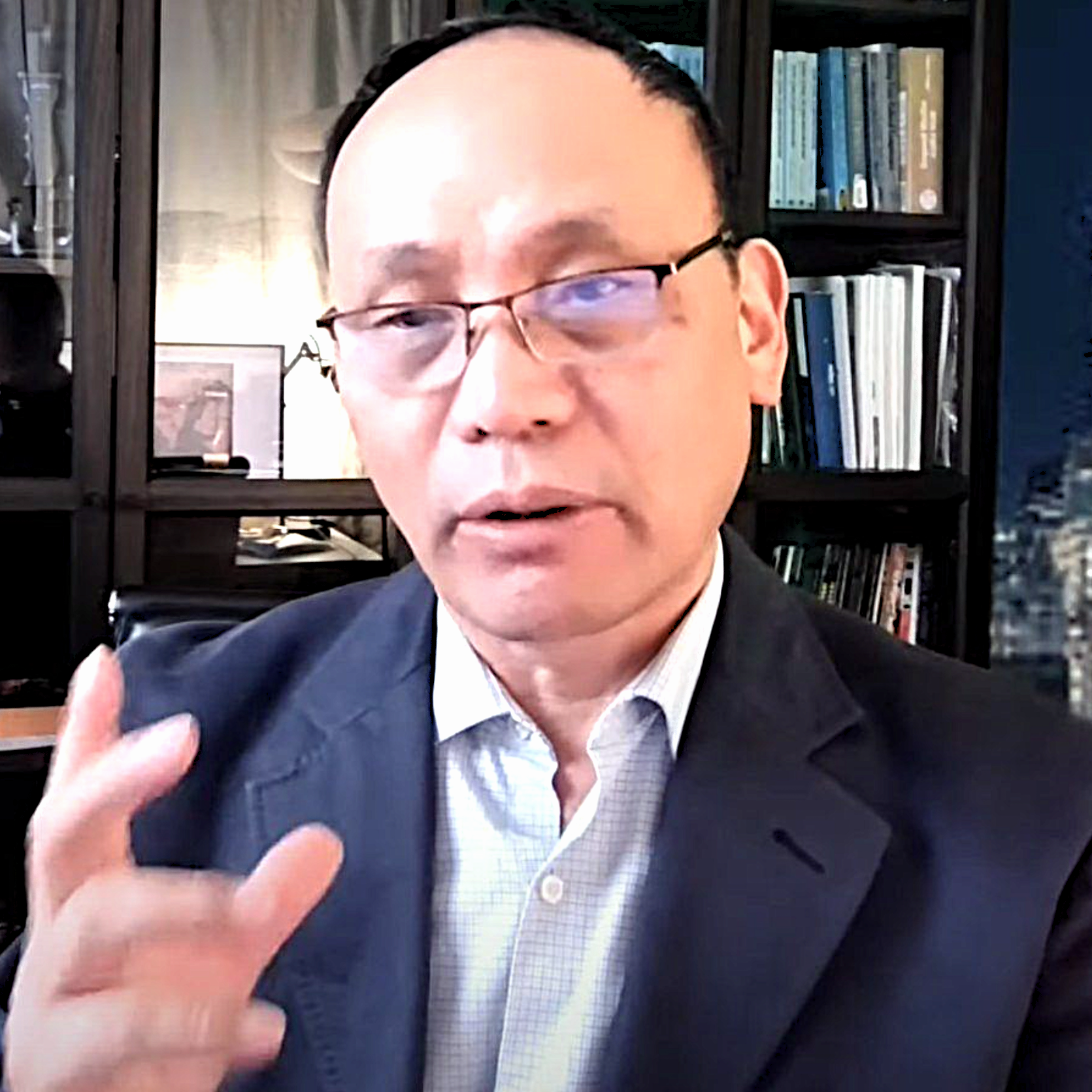 Headshot of Marohang Limbu wearing glasses, a light blue checkered dress shirt, and a dark navy blazer. He is pictured in front of a dark bookshelf filled with books.