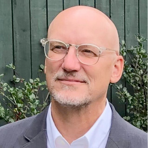 Professional headshot of Peter B. Hoffman wearing clear-framed glasses and a gray blazer over a white dress shirt. He is pictured outdoors in front of a dark green wooden fence.