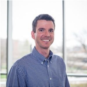 Professional headshot of Matthew Rynbrandt smiling, wearing a blue and white gingham checkered button-down shirt.