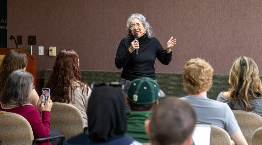 Cristina Rivera Garza stands at the front of a smaller room, holding a microphone and raising one hand as she speaks during the student-centered session. Students fill the rows of chairs facing her, some holding phones to capture the moment
