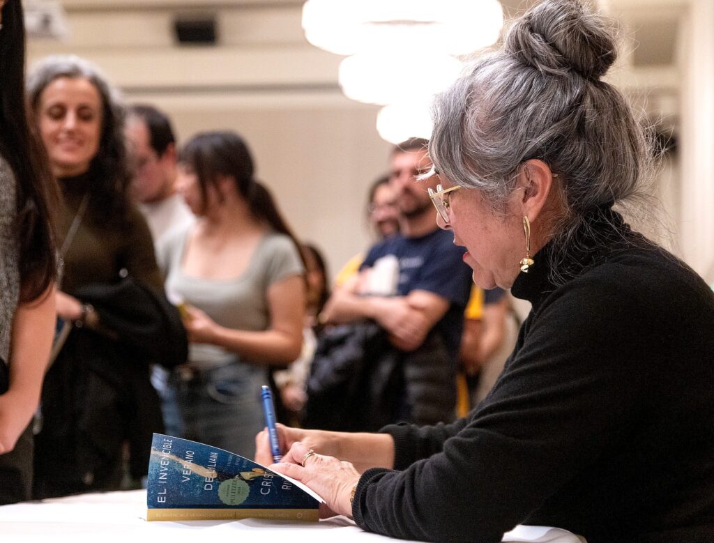 Cristina Rivera Garza bends over a table under warm overhead lighting, carefully signing a copy of the Spanish-language edition of her book, El Invencible Verano de Liliana, as a crowd of attendees waits in line behind her.
