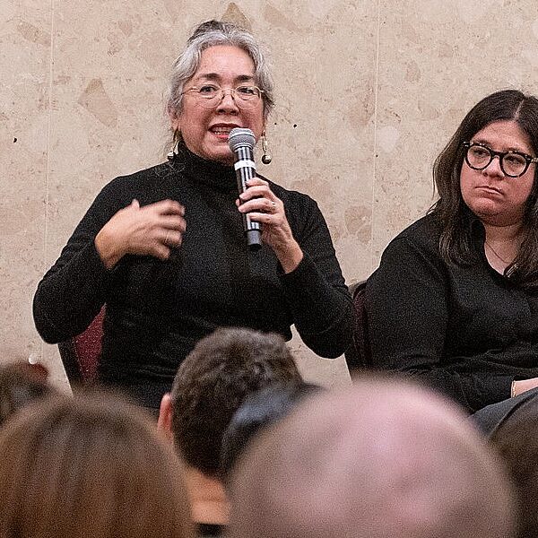 Cristina Rivera Garza speaks into a microphone at the left end of a faculty panel, flanked by another MSU faculty members seated before a full audience. A small high-top table with water bottles sits at the center of the panel arrangement.