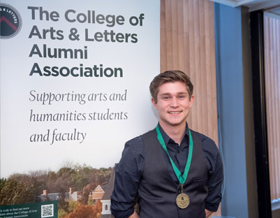 man with brown hair wearing medal posing next to "The College of Arts & Letters Alumni Association" sign