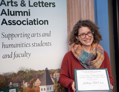 woman with glasses and scarf holding award posing next to "The College of Arts & Letters Alumni Association" sign