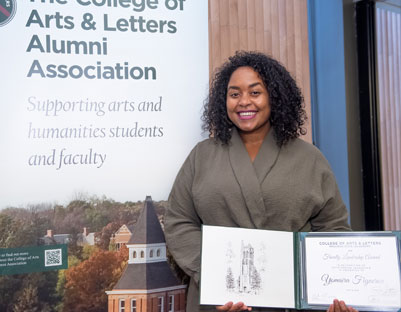 woman with black hair holding award posing next to "The College of Arts & Letters Alumni Association" sign