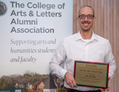 man with glasses holds award posing next to "The College of Arts & Letters Alumni Association" sign