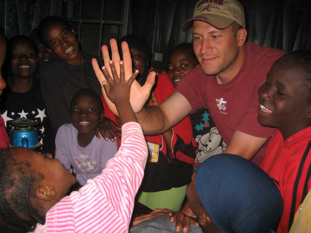 man in baseball cap surrounded by children high fiving child in kenya 