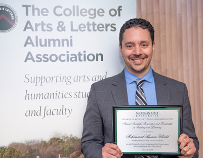 man with brown hair holding award posing next to "The College of Arts & Letters Alumni Association" sign