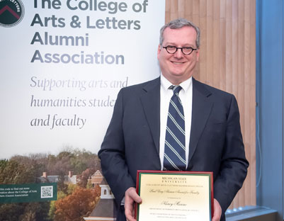 man with glasses holding award posing next to "The College of Arts & Letters Alumni Association" sign