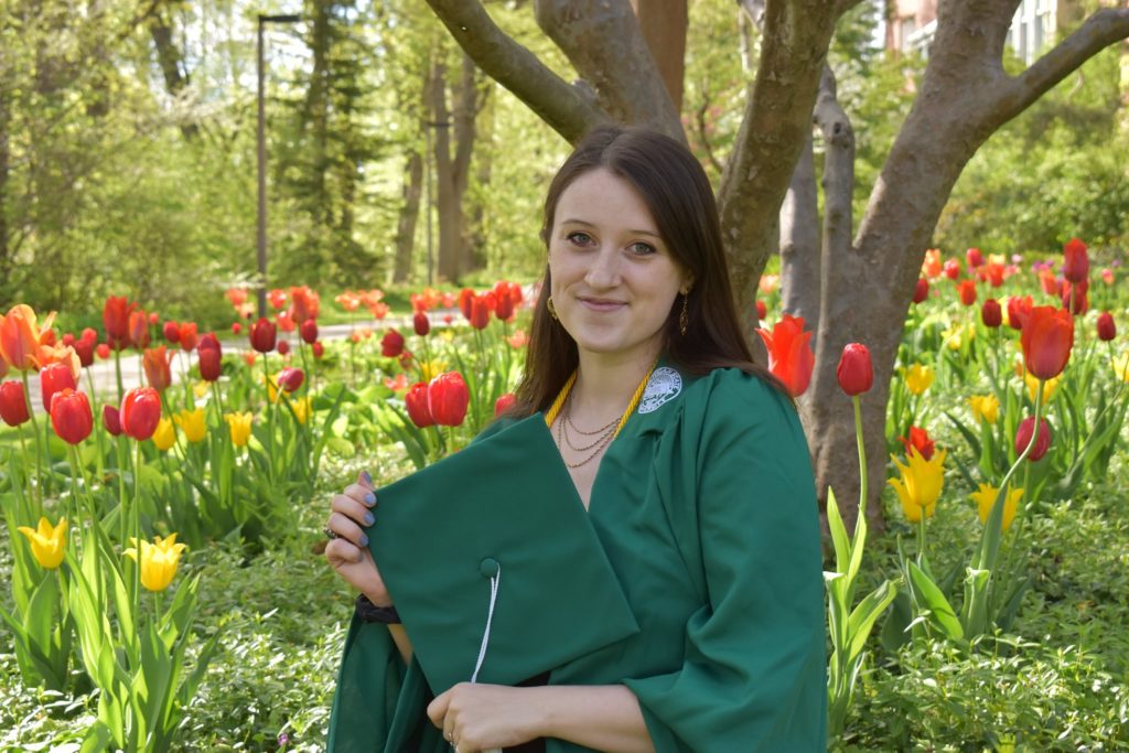 Photo of a woman with brown hair wearing a green graduation gown and holding a graduation cap in front of her chest. In the background are red and yellow tulips.