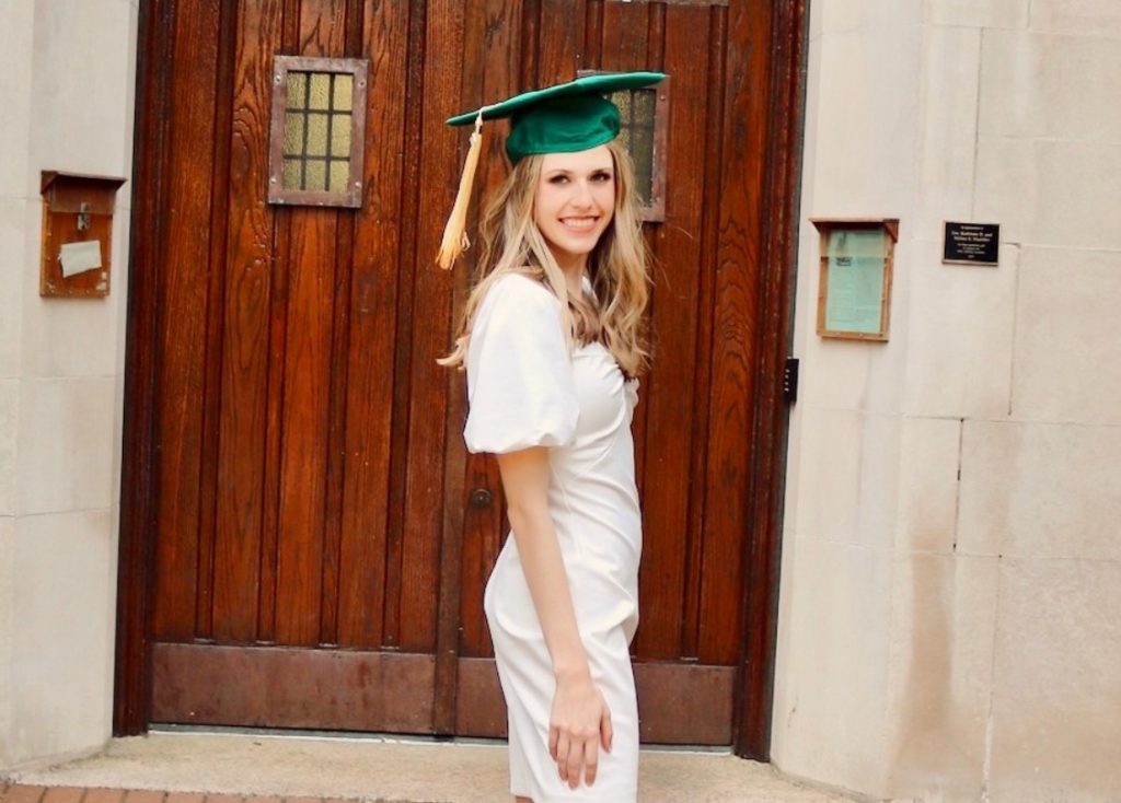 A girl with dark blonde hair wearing a white dress and a green graduation cap with a gold tassel smiling at the camera.