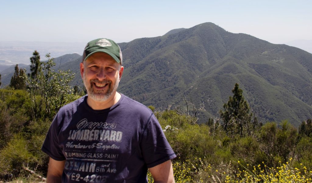 A picture of a man in a green hat and blue t-shirt smiling; there are mountains in the background.