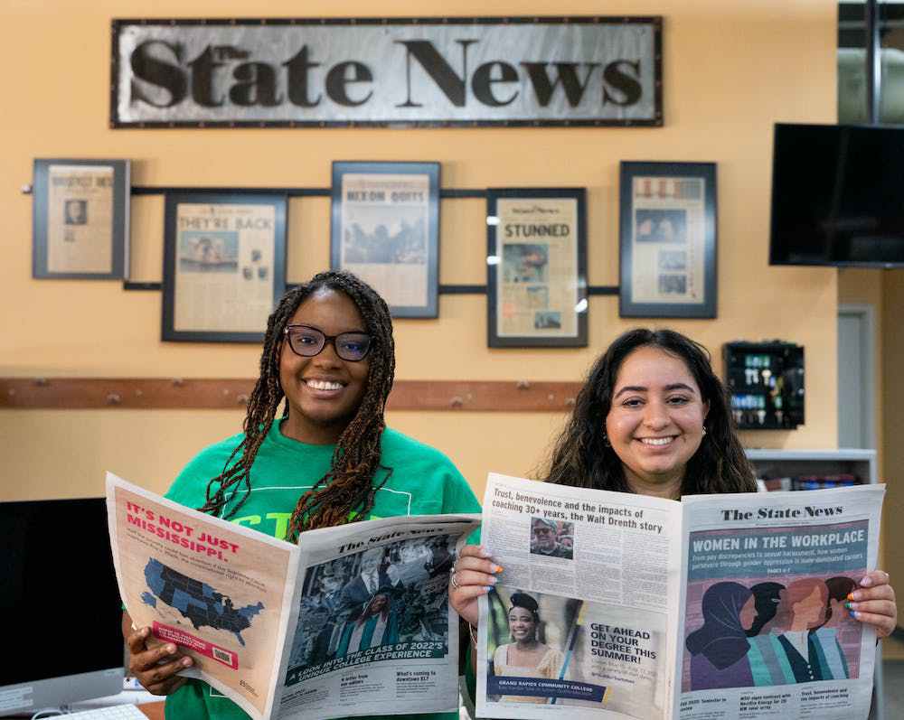 Two people smiling and looking at camera holding up a newspaper that reads, “The State News”.