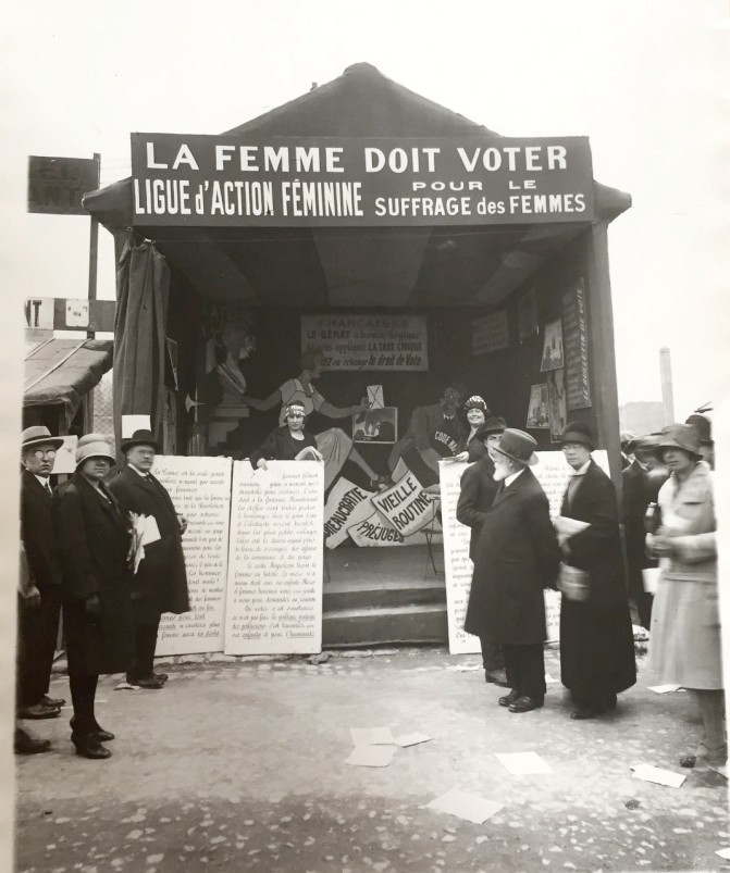 An old, black-and-white photograph of a feminist booth with an overhead banner reading, "THE WOMAN MUST VOTE". Other men and women gather in front of it.