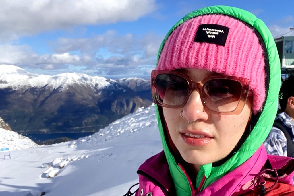 Woman wearing a pink knit hat and sunglasses with snow-capped mountains in the background. 