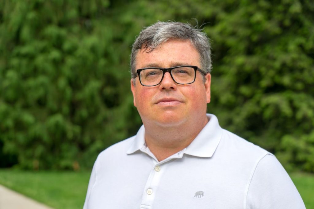 An outdoor portrait of a man with dark, graying hair wearing glasses and a white collared shirt.