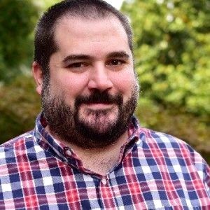 Portrait of a man with dark hair and beard wearing a red and blue plaid button-down.