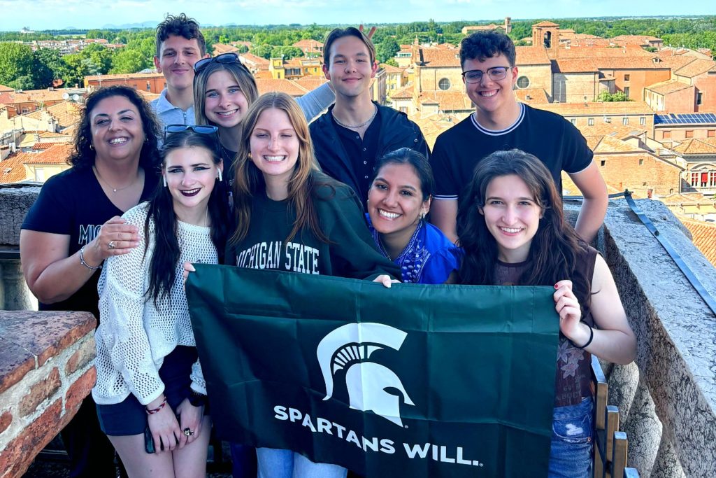 A group of nine individuals posing with a green flag with a Spartan helmet on it, above the text "Spartans Will." The background is a older Italian city with tan brick houses. 