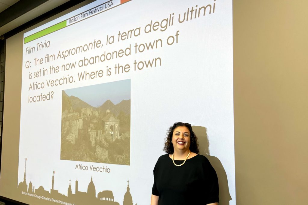 A picture of a woman with a black top and a pearl necklace standing in front of a large projector screen displaying a trivia question from the "Italian Film Festival USA." The slide reads, "Film Trivia: Q: The film Aspromonte, la terra degli ultimi is set in the now abandoned town of Africo Vecchio. Where is the town located?" Below the text is an image of the abandoned town with the label "Africo Vecchio."