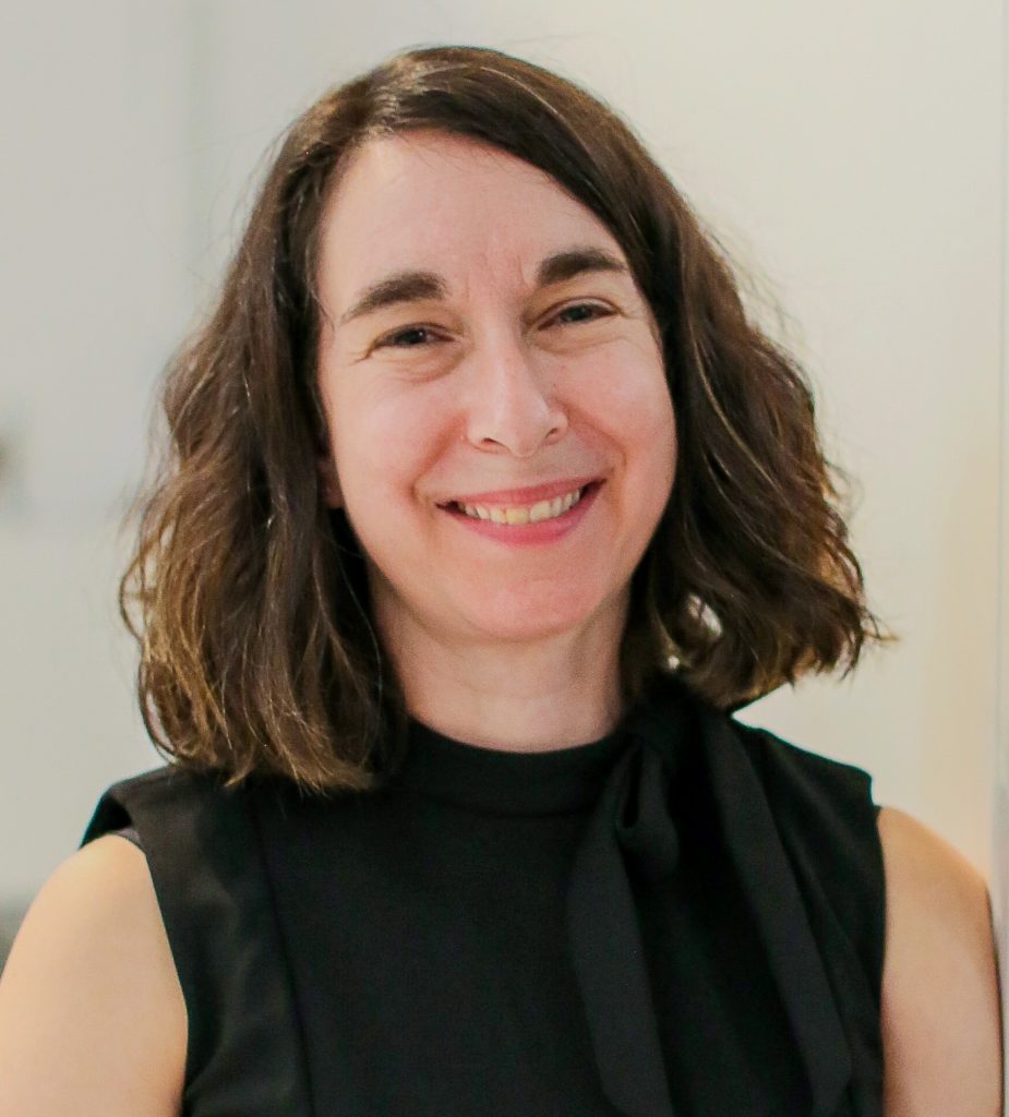 A picture of a woman in a black blouse with long brown hair smiling and posed in front of a white wall.