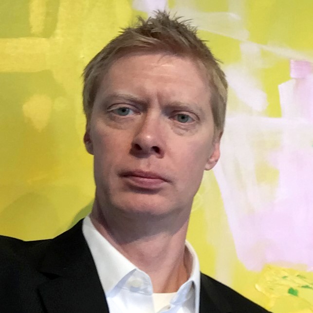 A picture of a man with short hair in a white button-down and black suit coat in front of a green and white wall. 